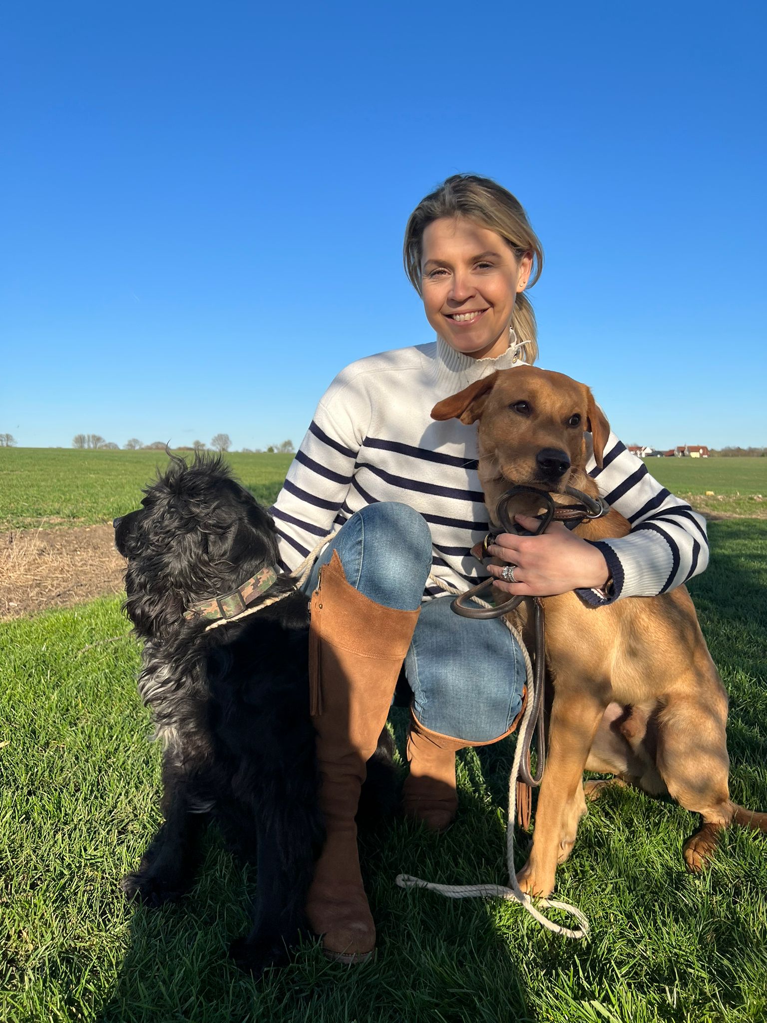 A woman kneels on the grass, smiling while holding two dogs on leashes. One dog is black and fluffy, and the other is brown with smooth fur. A clear blue sky and green fields are in the background.