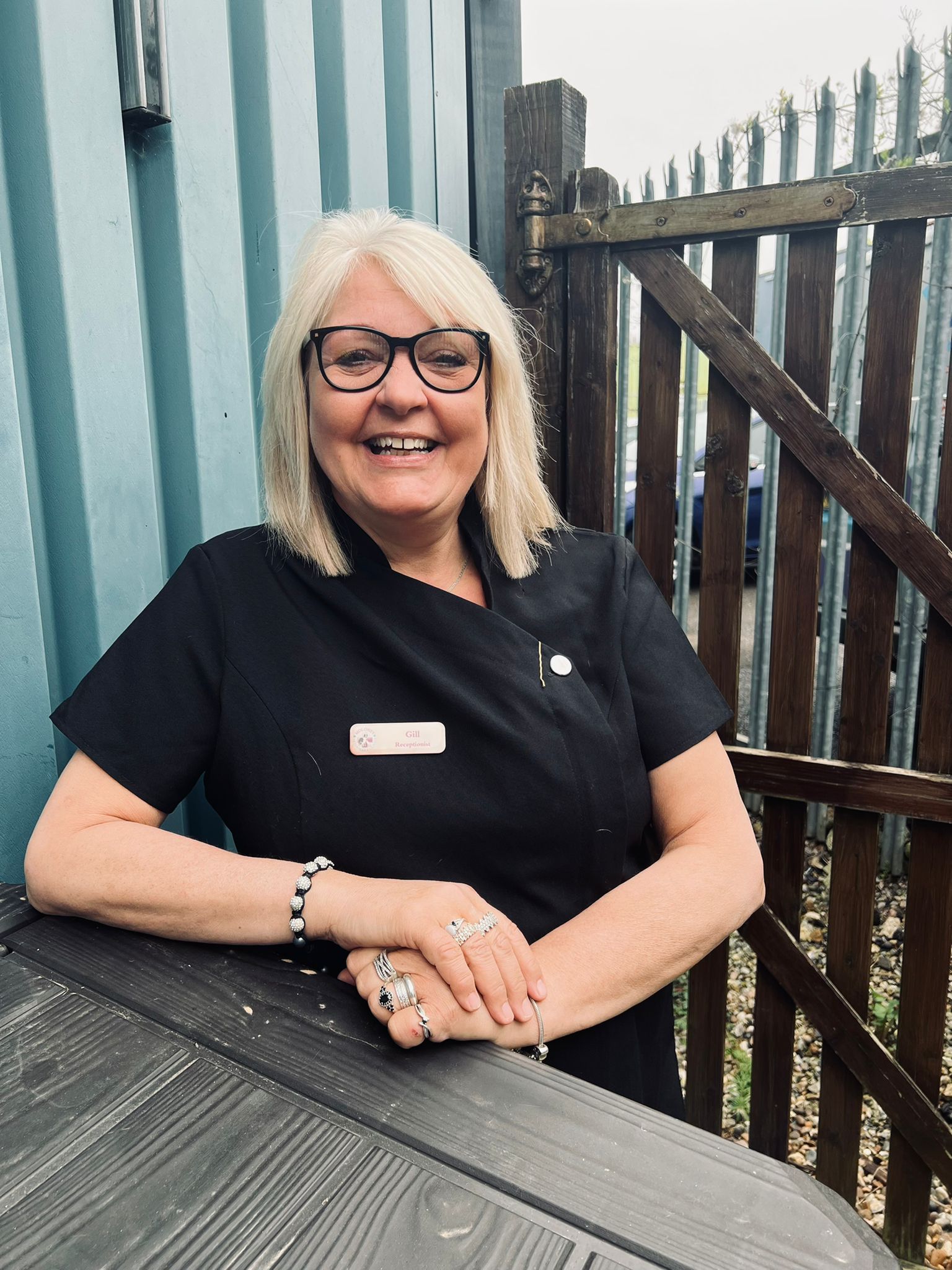 A smiling woman with blonde hair and glasses stands outside next to a wooden gate. She is wearing a black dress with a name badge and has her hands resting on a dark wood surface.