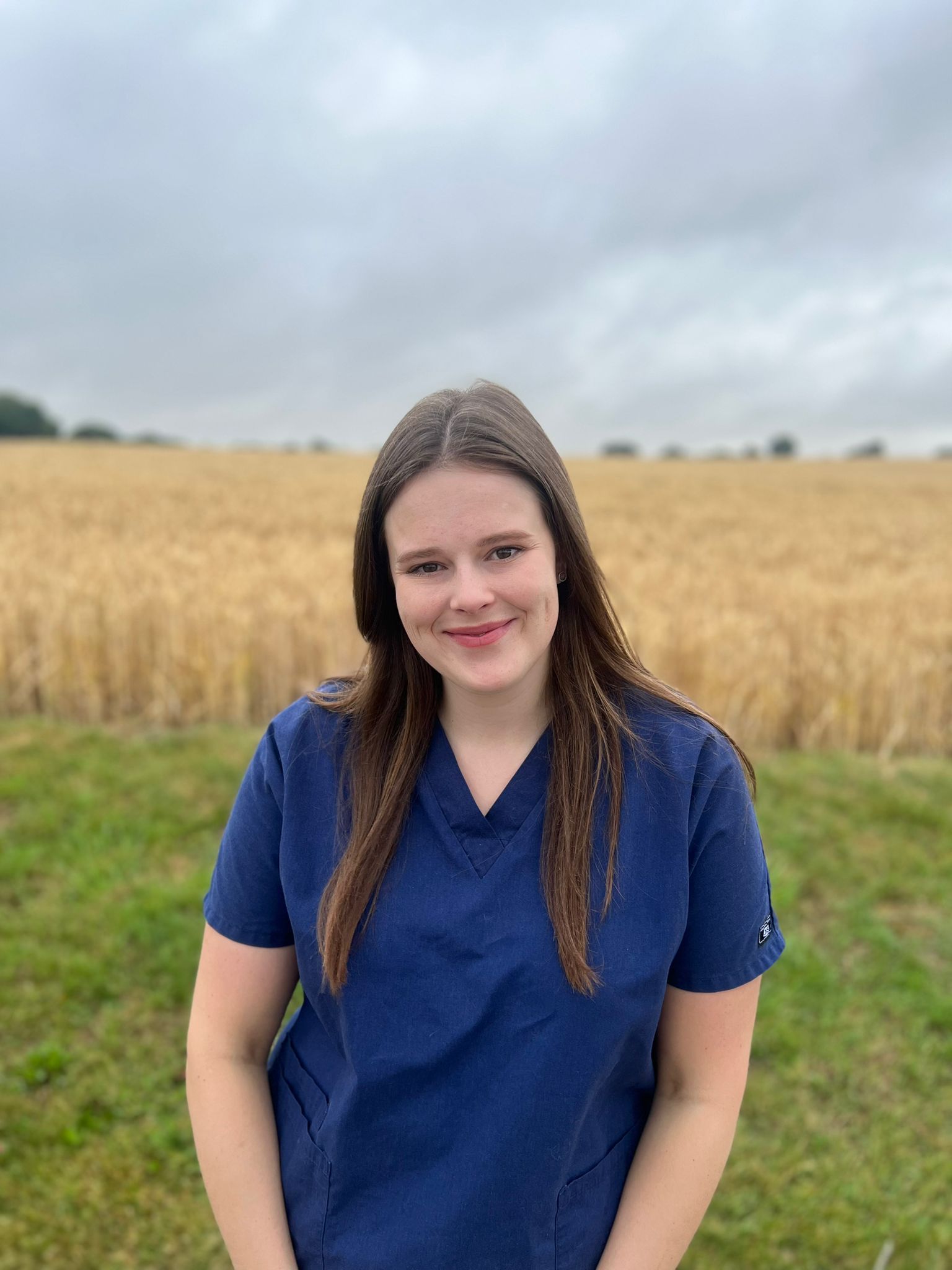 A woman with long brown hair, wearing a blue scrub top, stands smiling in front of a golden wheat field under a cloudy sky.
