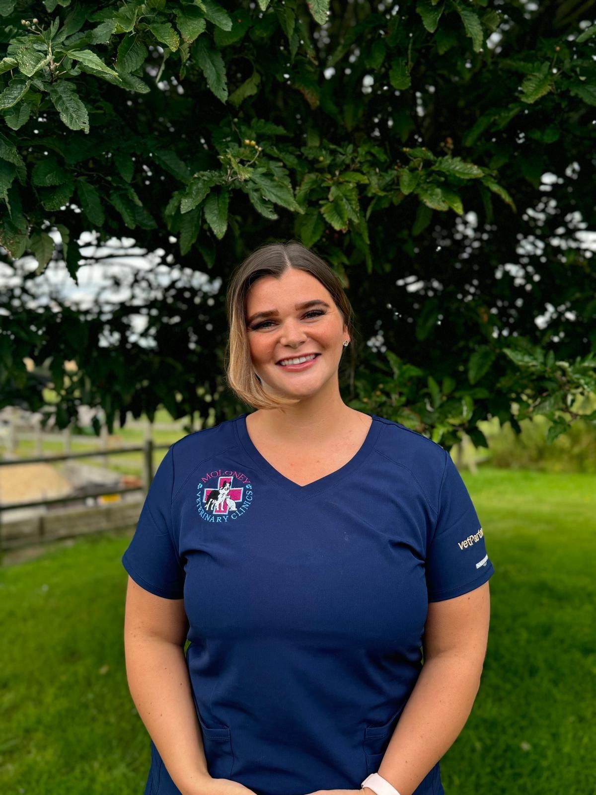 A person wearing a navy blue scrub top is smiling while standing outdoors in front of a lush green tree. The background features a grassy area and a wooden fence.