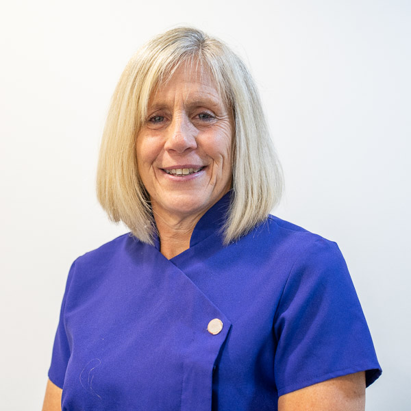 A woman with shoulder-length blonde hair is smiling, wearing a short-sleeved blue shirt. The background is plain white.