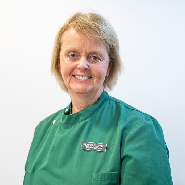 A person wearing a green uniform with a name tag smiles at the camera. The background is a plain light color.
