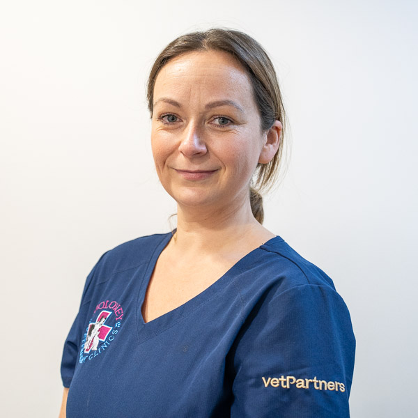 A person wearing a blue vetPartners scrub top smiles at the camera. The scrub features embroidered logos on the chest and sleeve. The background is plain and light-colored.