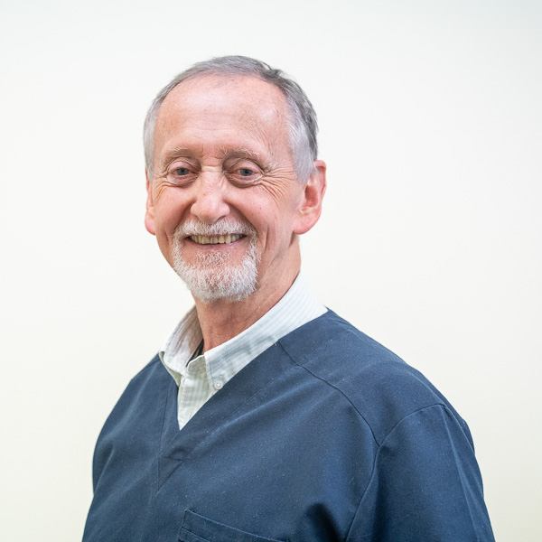 An older man with short gray hair and a beard smiles warmly. He wears a dark blue shirt over a collared white shirt. The background is a plain light color.