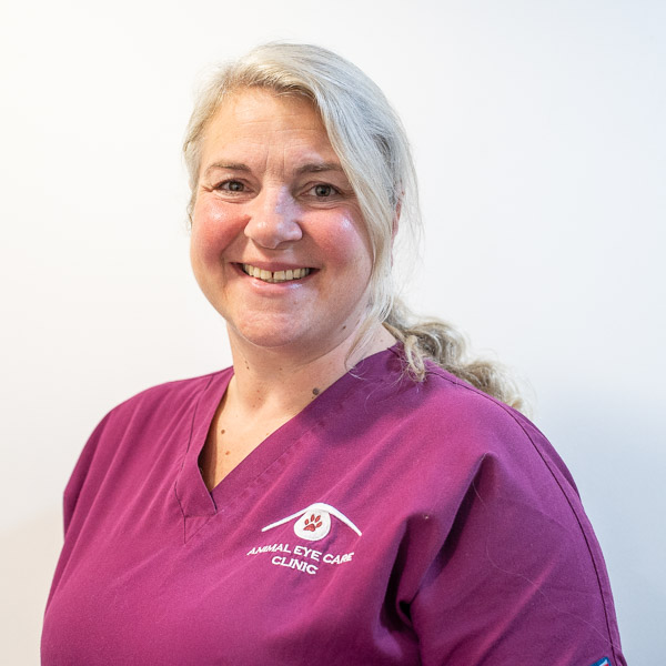 A person with long blonde hair is smiling at the camera. They are wearing a magenta scrub top with a logo that reads Animal Eye Care Clinic. The background is plain white.
