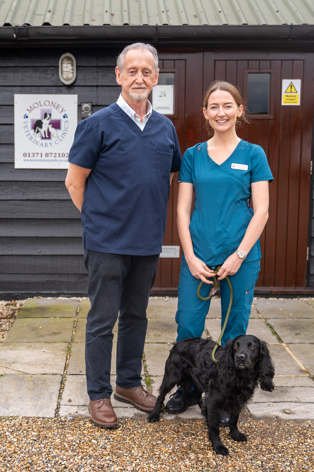 Two veterinary professionals, one man and one woman, stand smiling outside a clinic. The woman is holding a black dog on a leash. They are in front of a dark building with a sign indicating its a veterinary practice.