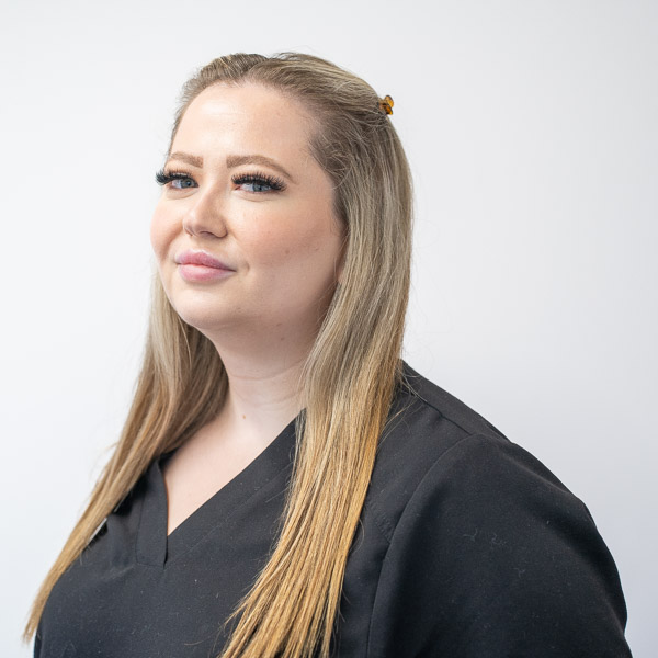 A woman with long blonde hair and a confident smile is wearing a black top. She stands against a plain white background, looking at the camera. Her hair is partially tied back.