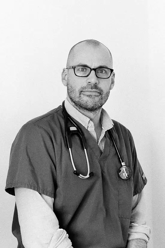 A man with glasses and a beard, wearing medical scrubs and a stethoscope around his neck, is looking at the camera. The photo is in black and white.